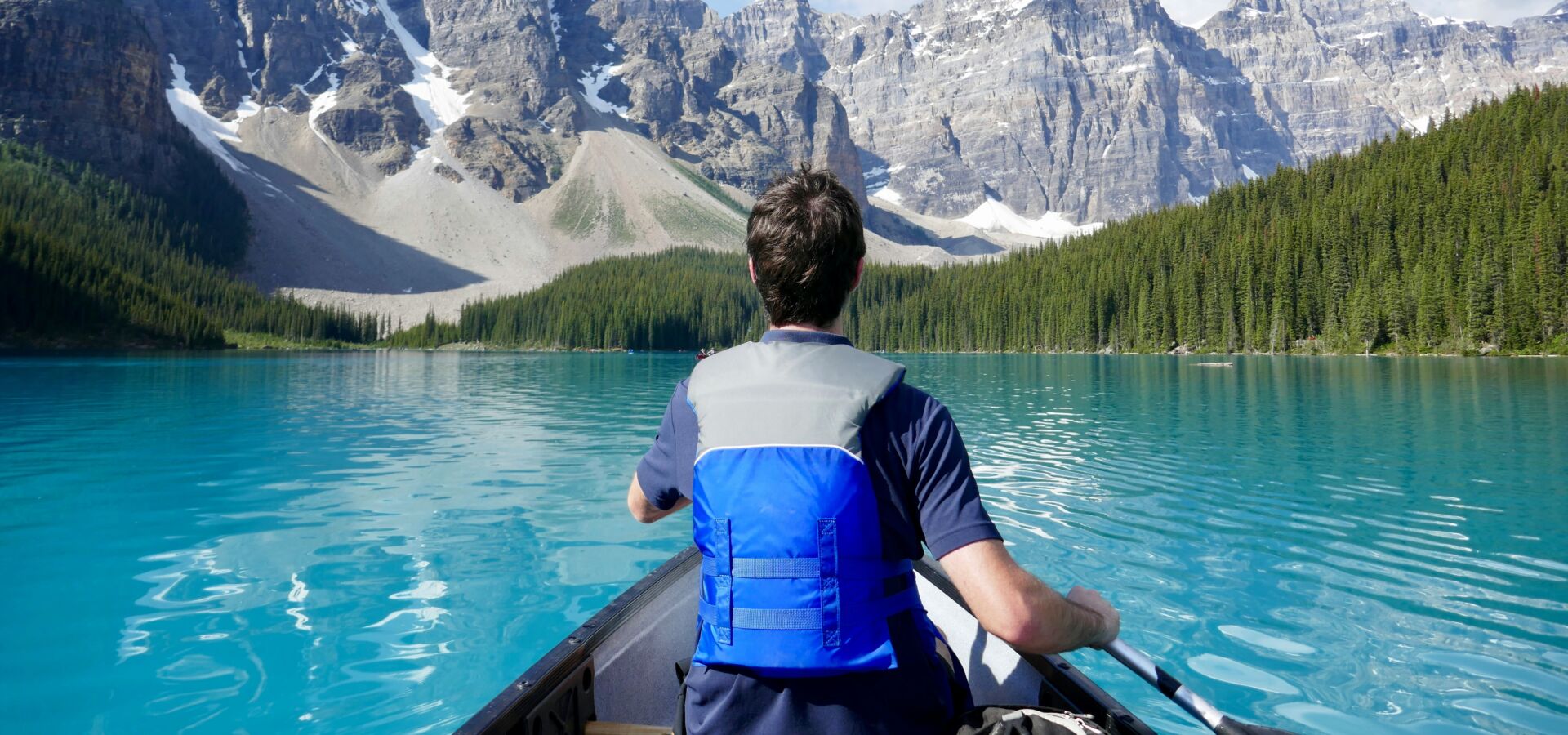 Mann sitzt in einem Kanu und fährt auf einem See. Im Hintergrund sieht man Berge.