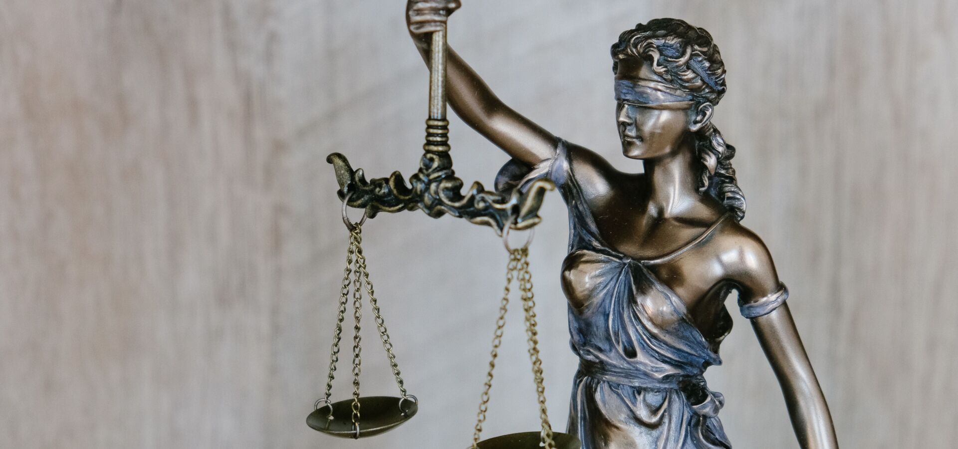Bronzefarbene Statue der Justitia mit Waage und Augenbinde als Symbol für Gerechtigkeit, Rechtsprechung und Unparteilichkeit vor hellem Hintergrund.